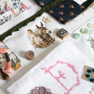 May include: A collection of craft supplies including floral fabric, green trim, embroidery floss in peach and brown, a wooden number 39 token, a small donkey and child sticker, a bird nest print, various buttons, and a white cloth with pink cross-stitch.