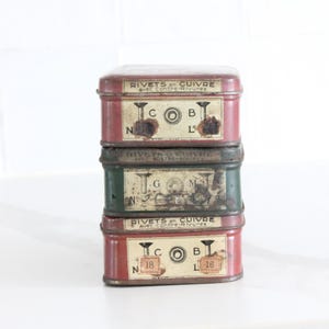 Peut inclure: Une pile de trois boîtes métalliques rectangulaires pour rivets en cuivre. Les boîtes du haut et du bas sont rouges avec des étiquettes crème portant l'inscription RIVETS en CUIVRE. La boîte centrale est verte avec une étiquette usée.