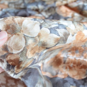 May include: Close-up of floral fabric featuring a pattern of large, abstract flowers in shades of brown, beige, and grey. The fabric is held in a hand, showing its texture and drape. The overall theme is textile design.