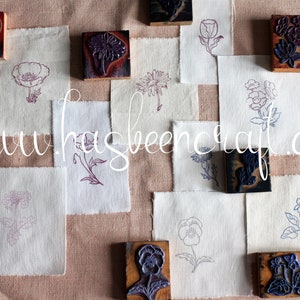 Op de afbeelding: Een verzameling houten stempels met bloemmotieven, gerangschikt op een beige stoffen achtergrond. De stempels zijn gesneden met verschillende bloemen, waaronder klaprozen, viooltjes en andere botanische motieven. De stof is textuurrijk en heeft een subtiel patroon. De tekst "www.hasbeencraft.com" is zichtbaar in het midden van de afbeelding.