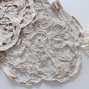 Peut inclure: Gros plan sur deux napperons en dentelle beige avec des motifs floraux complexes. Une couronne florale blanche est placée à droite des napperons. Les napperons ont un design ajouré délicat, parfait pour la décoration intérieure.