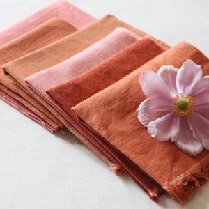 Op de afbeelding: Een stapel van zes vierkante stoffen in tinten oranje, roze en rood, met een enkele roze bloem bovenop. De stoffen vierkanten hebben een gefranst randje.