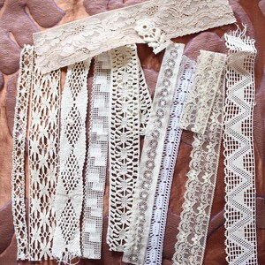 May include: Assortment of white lace trim, featuring various patterns and textures. The lace is arranged in a vertical line on a brown background.