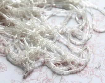 May include: A pile of clear glass beads, resembling small, rectangular tubes, arranged in a tangled heap. The beads are strung together on a thin thread, creating a delicate and intricate pattern.