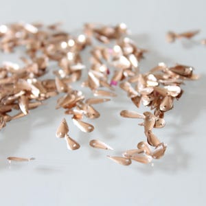 May include: A collection of small, metallic rose gold sequins shaped like teardrops. Each individual sequin features a single hole at the narrow end for attachment, with a smooth, reflective surface that catches light across the scattered arrangement.