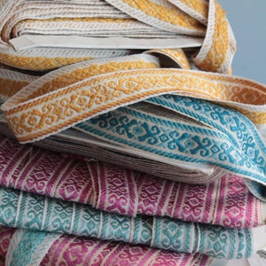 Puede incluir: Una pila de ribetes de tela coloridos con intrincados patrones. El ribete está en tonos dorados, azul verdoso, rosa y naranja. El ribete es perfecto para agregar un toque decorativo a la ropa, la decoración del hogar o las manualidades.