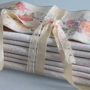 Op de afbeelding: Een stapel crèmekleurige stoffen stukken vastgebonden met een crèmekleurig lint en kant. Een bloemenrand met roze en gele rozen en andere bloemen ligt bovenop de stapel. Het lint is in een strik geknoopt.