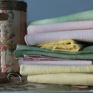 Peut inclure: Une bobine en bois avec du fil rose est à côté d'une pile de tissu aux couleurs pastel. Le tissu comprend des nuances de vert, de rose et de jaune. Un récipient décoratif avec des motifs floraux est également dans l'image.