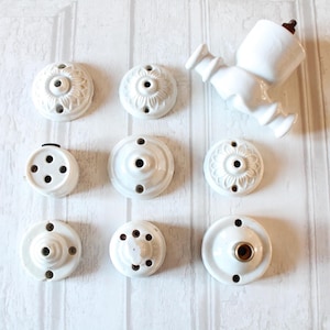 May include: A collection of white ceramic electrical fixtures, including ceiling roses and a light fixture. The ceiling roses have various floral and geometric designs. The light fixture is a vintage style. The items are arranged on a white wooden surface.