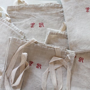 Peut inclure: Plusieurs sacs fourre-tout en lin beige, brodés d'initiales rouges. Les sacs, avec de longues anses, sont disposés en tas. Les initiales sont dans une police décorative. Le tissu est naturel et texturé.
