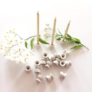 Peut inclure: Plusieurs petits bougeoirs en céramique blanc cassé avec une bougie dorée chacun. Des feuilles vertes et des fleurs blanches sont disposées autour des bougeoirs. D'autres bougeoirs sont éparpillés sur la surface blanche.