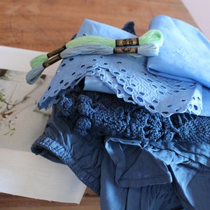 Op de afbeelding: Een verzameling blauwe stoffen en borduurgaren. Bevat een lichtblauwe broderie anglaise stof, een gevouwen blauwe doek en een donkerblauw gehaakt item. Twee spoelen groen en blauw borduurgaren liggen erop. Een print van een vrouw met bloemen staat links.