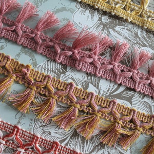 Può includere: Tre diversi passamanerie con frange, ciascuna in un colore diverso: rosa, oro e rosso. Le passamanerie sono realizzate in tessuto intrecciato con un motivo decorativo e hanno nappe sui bordi.