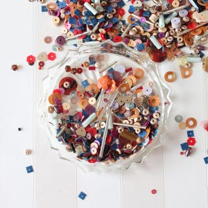 Op de afbeelding: Een doorzichtige glazen kom gevuld met een verscheidenheid aan losse knutselkralen en pailletten in diverse vormen, maten en kleuren, waaronder rood, blauw, oranje, bruin en metallic tinten. Extra losse kralen liggen verspreid rond de kom.
