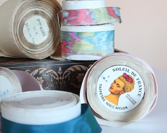 May include: Assortment of vintage ribbon spools in various colors, including cream, teal, and patterned designs. Some spools feature labels with text, including "SOLEIL DE FRANCE" and "100% NYLON".