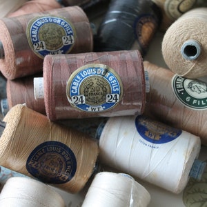 Puede incluir: Primer plano de una colección de carretes de hilo vintage en varios colores, incluyendo marrón, beige y blanco. Los carretes tienen etiquetas con el texto "CABLE LOUIS D'OR 6 FILS" y "THIR 6 FILS 24".