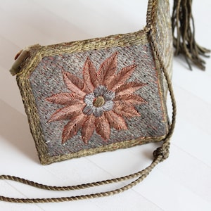 以下が含まれることがあります： 銅色と灰色の刺繍が施された長方形のテキスタイルアクセサリー。縁には太いメタリックゴールド調の編み込みトリムがあしらわれ、お揃いのツイストコードストラップと装飾的なタッセルが付いています。