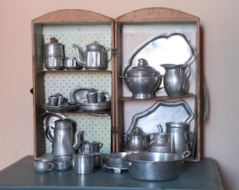 May include: A vintage miniature cabinet filled with antique silver-colored tea and coffee service pieces. The cabinet has two doors and is made of wood. The interior is decorated with a patterned wallpaper. The collection includes teapots, cups, saucers, and serving trays.