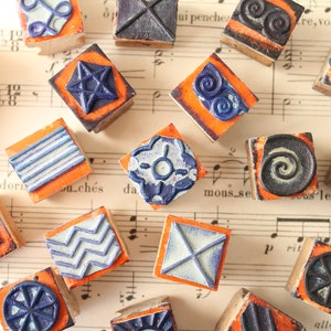 Può includere: Una collezione di 15 timbri in legno con vari disegni, tra cui forme geometriche, spirali e motivi astratti. I timbri sono dipinti in tonalità di blu e arancione.