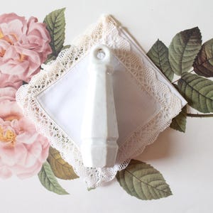 May include: A white ceramic object with a ring at the top, set on a white square doily with a lace edge. The doily is surrounded by a floral design of pink roses and green leaves, with text in the background.