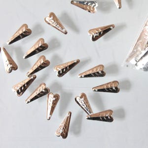 Op de afbeelding: Een verzameling kleine, zilverkleurige, driehoekige metalen onderdelen met een hartvormige bovenkant en drie horizontale gegraveerde lijnen. Deze metalen stukjes liggen verspreid, met een doorzichtig plastic zakje met meer onderdelen.