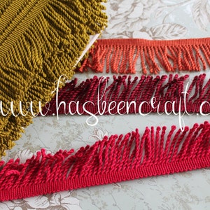 May include: Three different types of fringe trim in mustard yellow, coral orange, and crimson red. The trim is made of twisted cord and is approximately 1 inch wide.