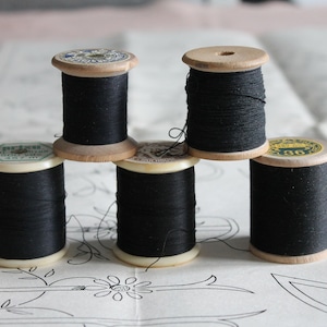 Op de afbeelding: Vijf vintage houten spoelen met zwart garen zijn gerangschikt op een oppervlak van papier met een patroon. De spoelen hebben een natuurlijke houten afwerking en zijn gevuld met strak gewikkeld zwart garen. Het papier heeft een bloemenpatroon.
