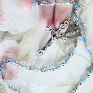 May include: A rosary featuring light blue faceted glass beads connected by silver-toned metal links. The rosary includes a silver-toned crucifix with a central sunburst design and a detailed oval-shaped metal centerpiece depicting a religious figure.