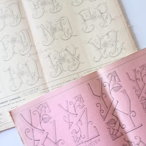Op de afbeelding: Oude open boeken met gedetailleerde, sierlijke letterontwerpen in zwarte inkt. Eén boek is lichtroze, de andere zijn gebroken wit. De letters zijn gestileerd met bloem- en decoratieve elementen, waarschijnlijk voor borduurwerk of design.
