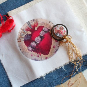 May include: A white fabric square with a printed image of a red heart with thorns and a crown of roses. A small round image of a child in a blue and white outfit is attached to a gold tassel.