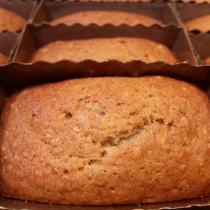 May include: Close-up of several golden-brown mini loaves of bread, each nestled in a brown cardboard container. The baked goods have a slightly textured surface and are arranged in neat rows, suggesting a bakery or homemade treat.
