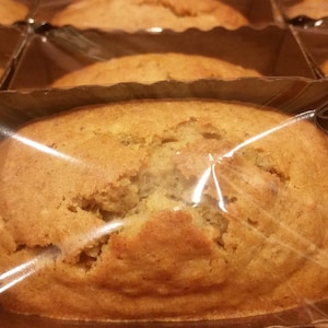 Puede incluir: Primer plano de varios panes dorados, envasados individualmente en film transparente y recipientes de cartón. El pan tiene una superficie ligeramente agrietada, lo que sugiere una calidad artesanal. La imagen está bien iluminada, destacando la textura y el color de los productos horneados.