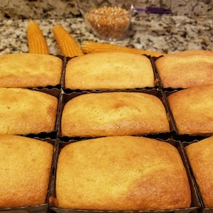 Puede incluir: Nueve panes de maíz dorados en moldes individuales. Los panes son rectangulares con una parte superior ligeramente redondeada. Mazorcas de maíz y un cuenco de granos de maíz en el fondo.