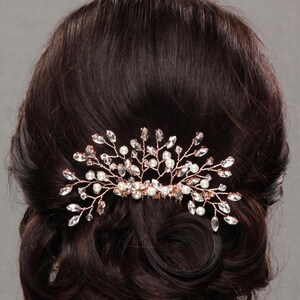 May include: A rose gold hair comb with a floral design, adorned with clear crystals and small white pearls. The comb is set against dark brown hair styled in an updo, suggesting bridal or formal wear.