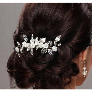 May include: A silver and white floral hair comb with clear crystals and white flowers. The comb is set in dark brown hair, with a pearl earring visible. The hair is styled in an updo.