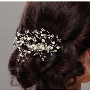 May include: A silver-toned hair comb with clear crystals and faux pearls. The comb is designed in a floral pattern and is set in dark brown hair. A pearl and crystal earring is visible.