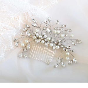 May include: A silver-toned bridal hair comb featuring a delicate design of clear crystals and faux pearls. The comb has a series of thin, silver teeth for secure placement in the hair. The crystals are various shapes and sizes, adding sparkle.
