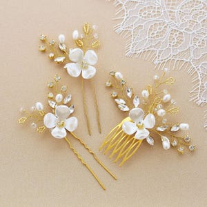 May include: Three gold-toned bridal hair accessories. The set includes two hair pins and a comb, all adorned with white flower accents, faux pearls, and clear crystals. The accessories are arranged on a neutral-toned surface.