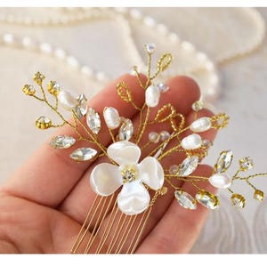 May include: A gold-toned hair comb featuring a central white flower with a crystal center, surrounded by pearl and crystal accents. Delicate gold wire branches extend from the flower, adorned with more crystals and pearls. The comb's teeth are also gold.