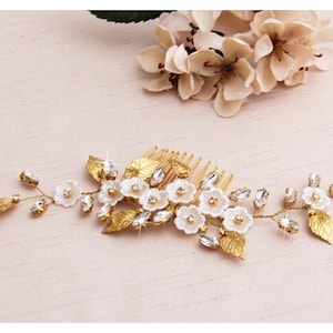 May include: A gold-toned hair comb adorned with white floral accents, clear crystals, and gold-colored leaves. The comb features a series of small, delicate flowers and sparkling crystals, creating an elegant and sophisticated accessory for special occasions.
