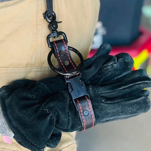 Op de afbeelding: Zwarte werkhandschoenen vastgemaakt met een zwarte leren riem en een metalen sluiting. De riem heeft rode stiksels en is bevestigd aan een zwarte karabijnhaak. De handschoenen zijn bevestigd aan een beige uniform.
