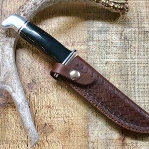 Könnte beinhalten: Eine braune Lederscheide mit einem gewebten Muster und einem Druckknopfverschluss. Ein Jagdmesser mit schwarzem Griff und silberner Klinge befindet sich in der Scheide.