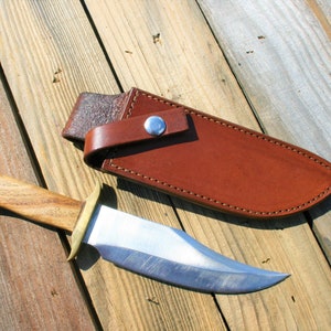 May include: A handmade hunting knife with a wooden handle and a polished steel blade, resting beside a brown leather sheath. The knife features a brass bolster and a full tang construction. The sheath has a belt loop and a snap closure.