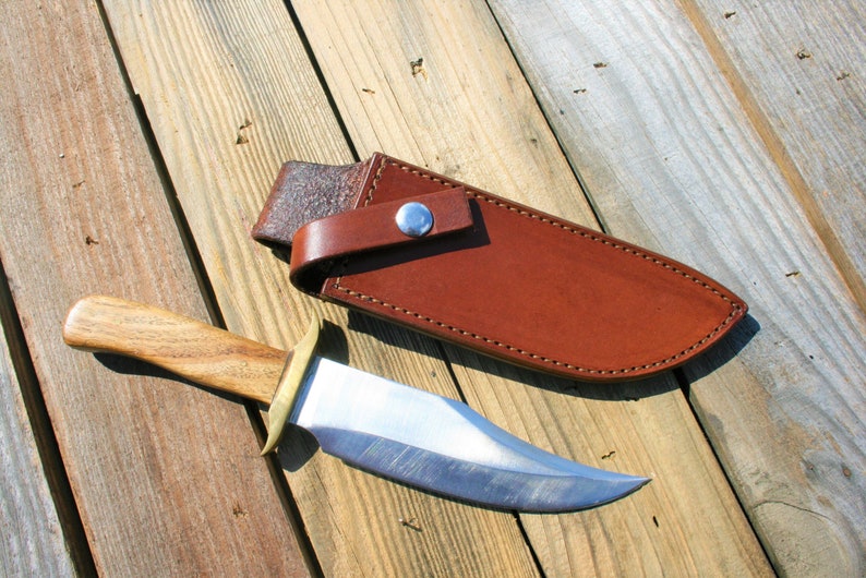 May include: A handmade hunting knife with a wooden handle and a polished steel blade, resting beside a brown leather sheath. The knife features a brass bolster and a full tang construction. The sheath has a belt loop and a snap closure.