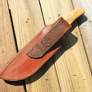 May include: A handcrafted knife with a wooden handle and a leather sheath. The sheath is brown with a stitched border and a strap with an embossed design. The knife is resting on a wooden surface.