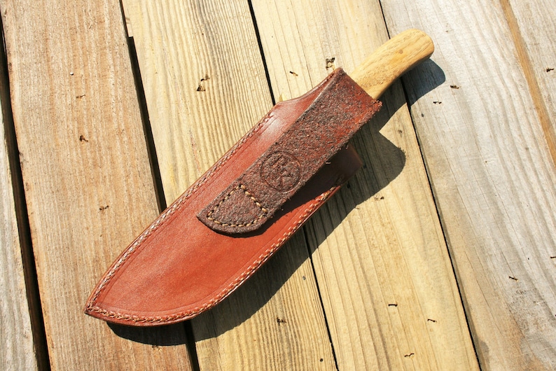 May include: A handcrafted knife with a wooden handle and a leather sheath. The sheath is brown with a stitched border and a strap with an embossed design. The knife is resting on a wooden surface.