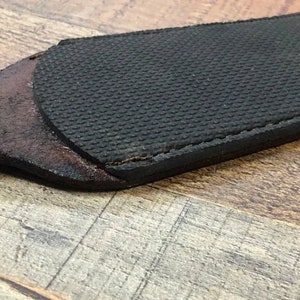 May include: A close-up of a brown leather strop with a black rubberized surface. The strop is used for sharpening knives.