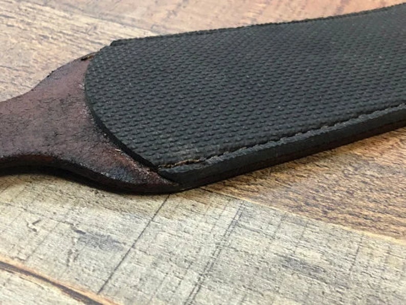 May include: A close-up of a brown leather strop with a black rubberized surface. The strop is used for sharpening knives.