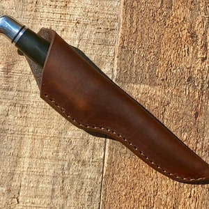 Puede incluir: Una funda de cuchillo de cuero marrón con un borde cosido. La funda está vacía y está sobre una superficie de madera.