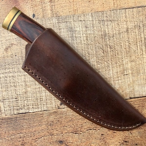 May include: A brown leather knife sheath with a stitched edge. The sheath is empty and has a brown wooden handle with a brass accent.